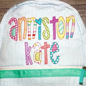 May include: A light blue and white striped backpack with a green zipper and a green ribbon. The backpack has the names "Anniston Kate" embroidered on the front in colorful patchwork letters.