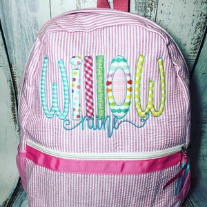 May include: Pink and white striped backpack with the name "Willow Craine" embroidered in colorful letters. The backpack has a pink handle, a white zipper, and a pink ribbon detail. A side pocket is visible.