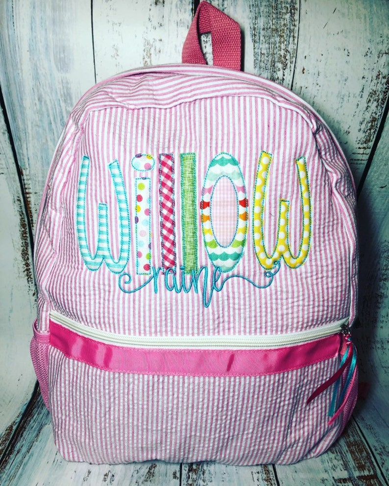May include: Pink and white striped backpack with the name "Willow Craine" embroidered in colorful letters. The backpack has a pink handle, a white zipper, and a pink ribbon detail. A side pocket is visible.