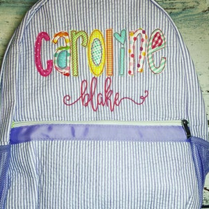 May include: A seersucker backpack with vertical stripes in white and light purple. The backpack features the name "Caroline" in colorful, embroidered letters, with "Blake" embroidered below. It has a zippered pocket and a mesh side pocket.