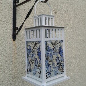 May include: A white metal lantern featuring glass panels hand-painted with blue birds and floral branches. The lantern has a square frame, a top handle, and is suspended from a black wrought iron wall bracket with a curved decorative design.