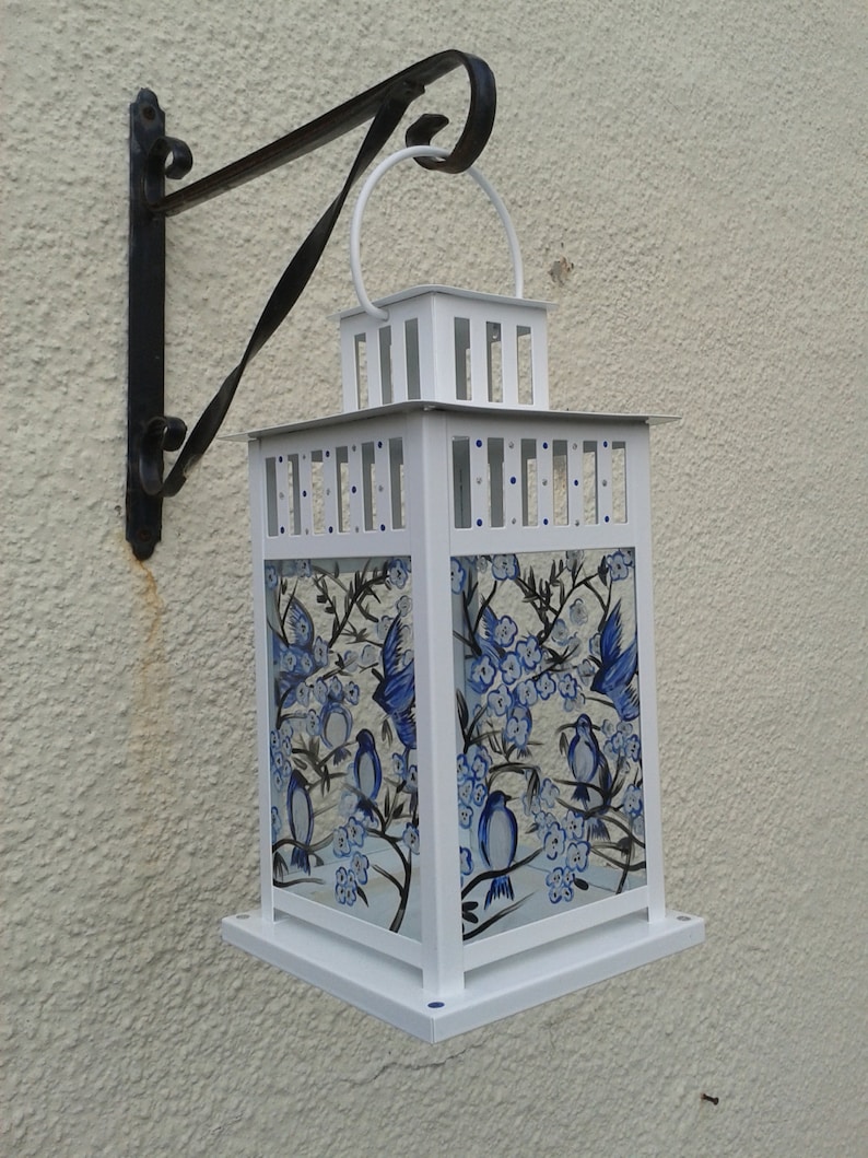 May include: A white metal lantern featuring glass panels hand-painted with blue birds and floral branches. The lantern has a square frame, a top handle, and is suspended from a black wrought iron wall bracket with a curved decorative design.