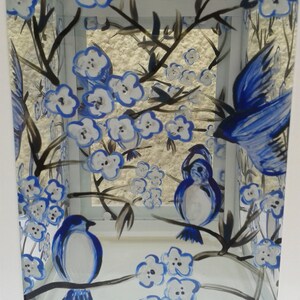 May include: A white metal lantern featuring hand-painted blue and white floral blossoms and blue birds perched on dark brown branches. The glass panels are decorated with a nature-inspired motif of birds in flight and resting on flowering stems.