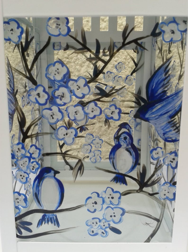 May include: A white metal lantern featuring hand-painted blue and white floral blossoms and blue birds perched on dark brown branches. The glass panels are decorated with a nature-inspired motif of birds in flight and resting on flowering stems.