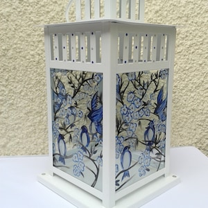 May include: A white metal lantern featuring glass panels hand-painted with blue birds perched on branches with blue blossoms. The lantern has a square base, a slatted top section with small blue and silver accents, and a white handle on top.