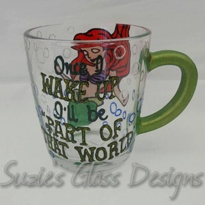 Puede incluir: Una taza de vidrio transparente con un asa verde, pintada a mano con una imagen de dibujos animados de Ariel de La Sirenita. La taza tiene el texto "Once I Wake Up I'll Be Part Of That World" pintado en ella.