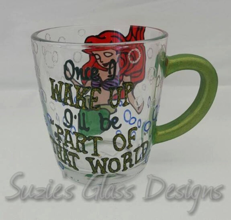Puede incluir: Una taza de vidrio transparente con un asa verde, pintada a mano con una imagen de dibujos animados de Ariel de La Sirenita. La taza tiene el texto "Once I Wake Up I'll Be Part Of That World" pintado en ella.