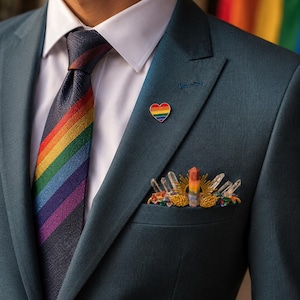 Op de afbeelding: Een donkerblauwgroen colbert gedragen met een wit overhemd en een diagonaal gestreepte regenboogdas. Een kleine hartvormige regenboogspeld is op de revers bevestigd en een pochet bevat een centrale regenboogkristal met kwartspunten.