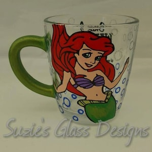 Puede incluir: Una taza de vidrio verde con una imagen pintada a mano de Ariel, la Sirenita, con cabello rojo, un sujetador de concha p&uacute;rpura y una cola verde. La taza tiene burbujas azules y blancas pintadas alrededor de la imagen.