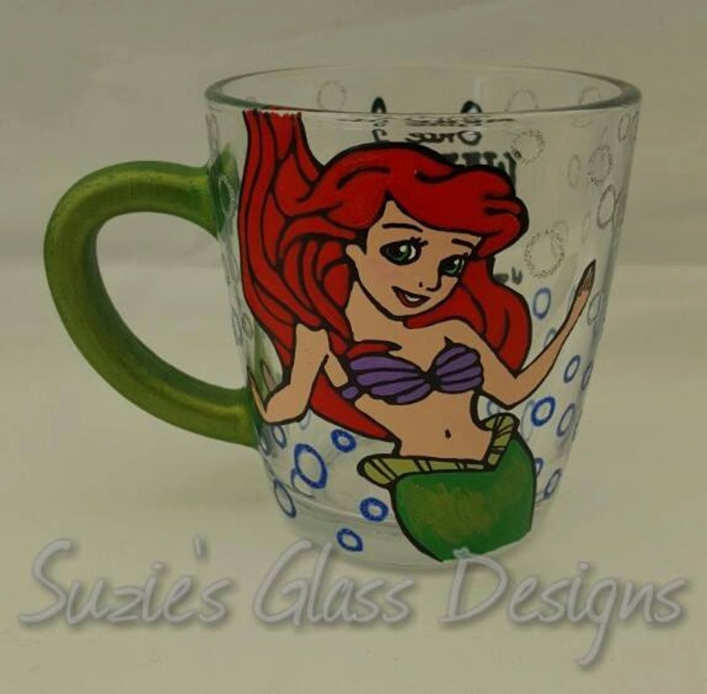 Puede incluir: Una taza de vidrio verde con una imagen pintada a mano de Ariel, la Sirenita, con cabello rojo, un sujetador de concha p&uacute;rpura y una cola verde. La taza tiene burbujas azules y blancas pintadas alrededor de la imagen.