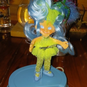May include: A vintage doll with blue hair and a green outfit stands on a blue base. The doll has a yellow face and blue shoes. The doll's hair is styled in a voluminous updo with green and blue accents. The doll is a collectible item.
