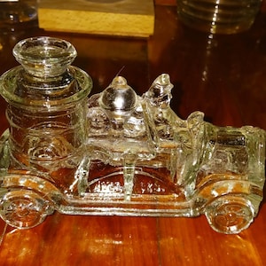 May include: A clear glass train-shaped container with a cylindrical body and a flat top. The container has a detailed design, including wheels and a small figure on top. The glass has a slight amber tint, and it sits on a wooden surface.