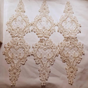 May include: Six off-white lace appliqués featuring intricate floral embroidery, a lattice mesh pattern, and small pearl bead accents. Each piece has a symmetrical, elongated shape with scalloped edges, designed for sewing onto fabric surfaces.