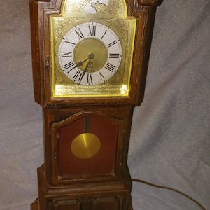 May include: A wooden mantel clock with a dark brown finish, featuring a gold-toned face with black Roman numerals and the text Tempus Fugit. The clock has a glass-fronted lower section displaying a circular gold-colored pendulum and a power cord.