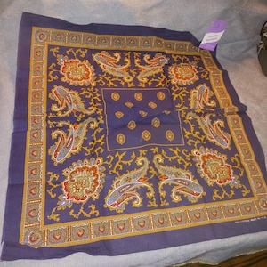 Puede incluir: Un pañuelo cuadrado azul marino con un detallado estampado de cachemira en rojo, amarillo y azul. El borde presenta un diseño floral y de corazón repetido. La etiqueta dice "Novelty Bandana".