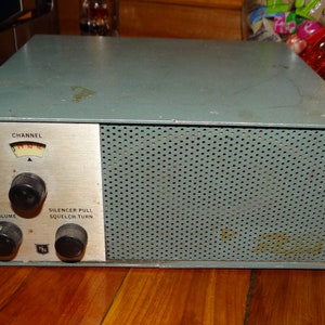 Puede incluir: Receptor de radio vintage de color verde azulado con un panel frontal perforado. Cuenta con un panel de control plateado con perillas para volumen, selección de canal y squelch. El indicador de canal muestra una aguja que apunta a una escala numerada.