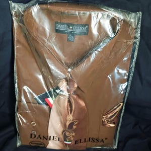 Puede incluir: Camisa marrón con corbata y pañuelo a juego, empaquetada en plástico transparente. La camisa tiene cuello puntiagudo y la marca "DANIEL ELLISSA NEW YORK" es visible en la etiqueta.