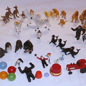 May include: A collection of small plastic animal figurines including elephants, polar bears, kangaroos, zebras, tigers, gorillas, and seals. Also included are a red tractor, a ringmaster figure, several colorful spheres, and a red top hat accessory.