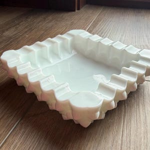 May include: A square, white ashtray with a scalloped edge. The ashtray is made of a solid, opaque material and sits on a wooden surface. The design is simple and elegant.