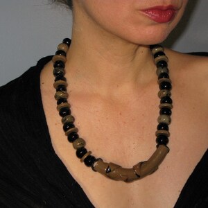 May include: A statement necklace featuring a mix of black and tan beads. The necklace has a curved, tan-colored centerpiece. The beads are round and vary in size, creating a bold and eye-catching accessory.