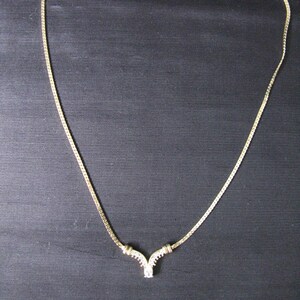 May include: A gold-toned necklace with a delicate chain and a V-shaped pendant. The pendant is adorned with small, clear gemstones. The necklace is displayed against a dark background.