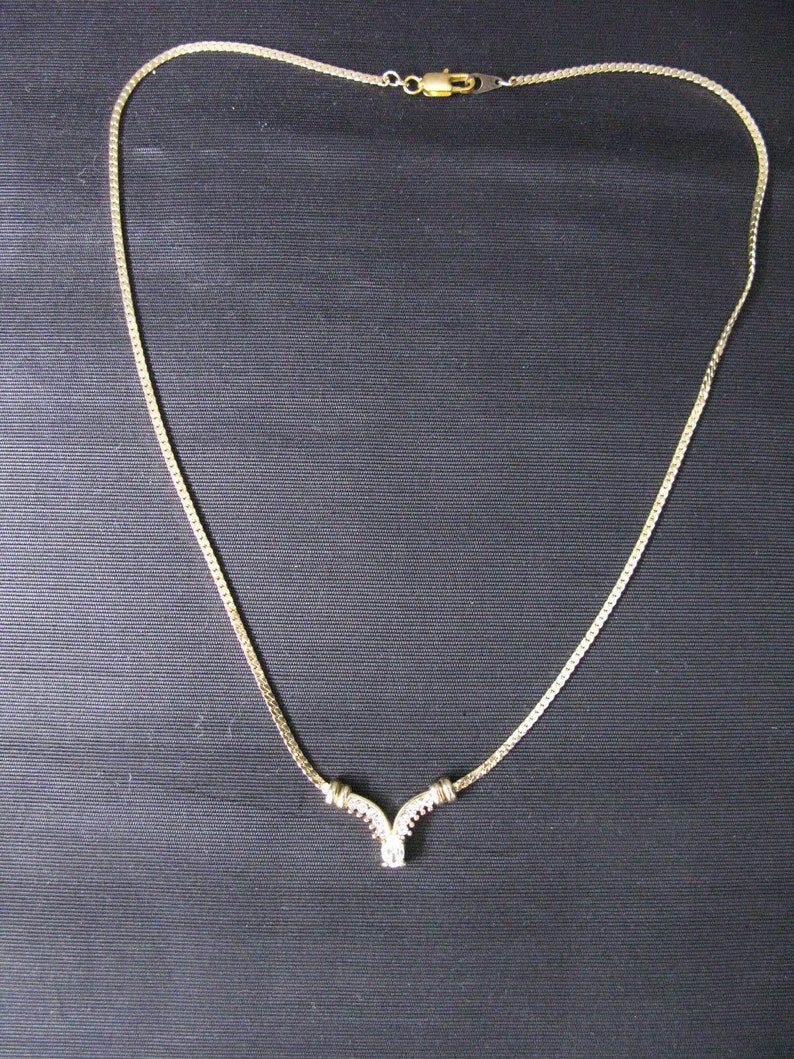 May include: A gold-toned necklace with a delicate chain and a V-shaped pendant. The pendant is adorned with small, clear gemstones. The necklace is displayed against a dark background.