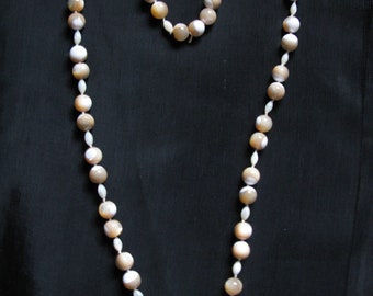 May include: A necklace composed of alternating round, light brown beads and smaller, white, oval-shaped beads. The necklace is arranged in a heart shape against a black background. The beads have a pearlescent sheen.