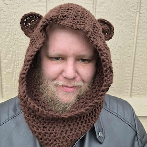 May include: A brown crocheted bear hood with attached ears. The hood is designed to cover the head and neck, creating a cozy and whimsical look. The hood is made of a textured yarn, and is a fun accessory for costumes or everyday wear.