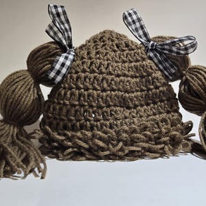 May include: A brown crocheted hat with two yarn pigtails and two black and white checkered bows. The hat is made of a textured yarn and is designed to resemble a doll's hairstyle.