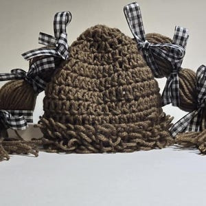 May include: A brown crocheted hat with attached yarn hair and three yarn ball accents. Each yarn ball is adorned with a black and white checkered ribbon bow. The hat and hair are made of a textured yarn.