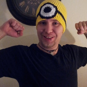 May include: A person wearing a yellow crocheted hat with a black and white eye design. The hat is similar to the character of a minion from the movie Despicable Me.