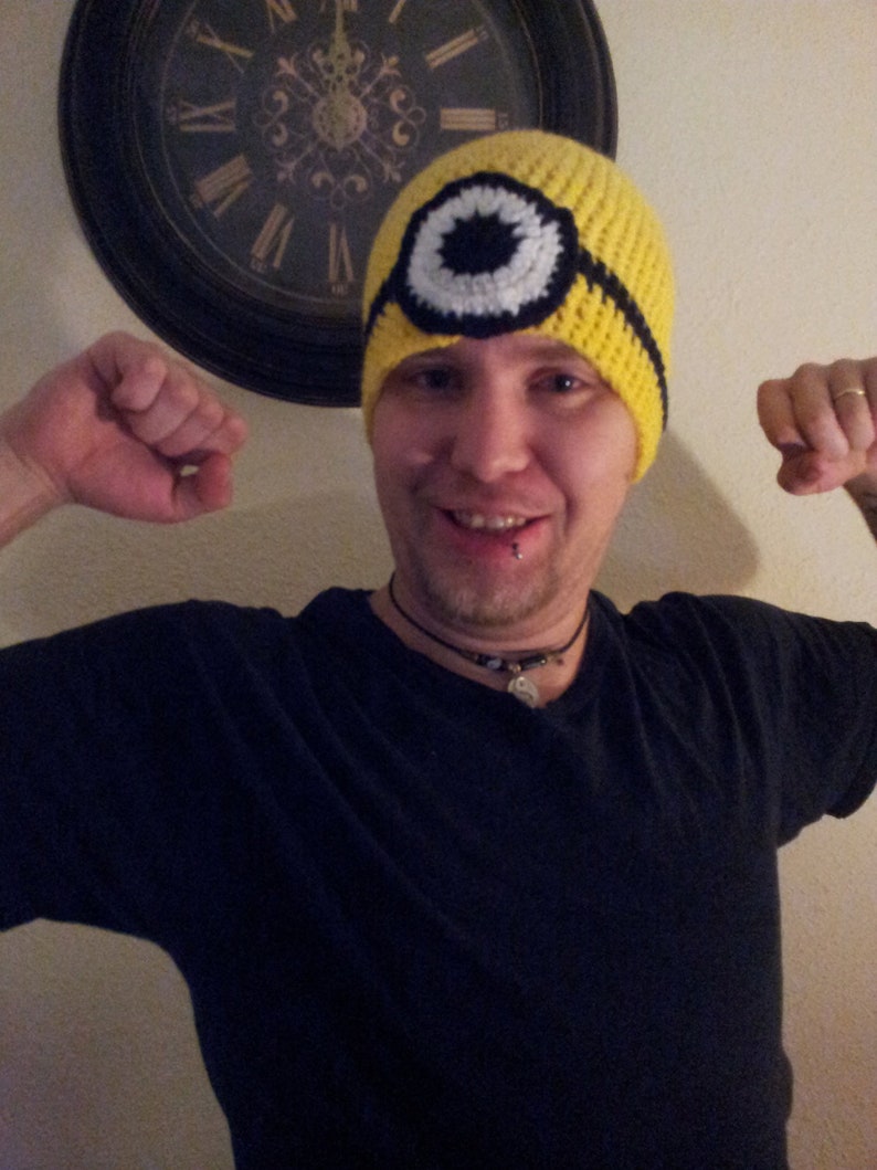 May include: A person wearing a yellow crocheted hat with a black and white eye design. The hat is similar to the character of a minion from the movie Despicable Me.