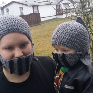 May include: Two gray crocheted knight helmets with attached face guards. The helmets have a textured pattern and a fin-like detail at the back. The face guards are striped in black and gray.
