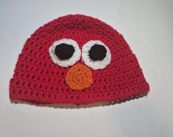 May include: A red crocheted hat with a character face design. The hat features white and black eyes and an orange nose. The hat is made of a soft, textured yarn and is likely for a child.