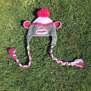 May include: A gray and white crocheted monkey hat with pink accents and a pink pom-pom on top. The hat has long, braided ties.