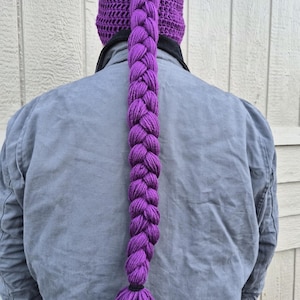 May include: A vibrant purple crocheted beanie featuring a long, thick, central braid that extends down the back. The hat is crafted with a textured stitch pattern and terminates in a matching purple yarn tassel at the bottom of the braid.