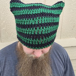 May include: A handmade crochet hat in alternating stripes of dark green and navy blue. The hat features two pointed ear-like protrusions at the top. The hat is made of a thick yarn, and the stitches are clearly visible.