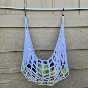 May include: A white, open-weave crochet bag filled with green and yellow fruit, hanging against a light brown wooden wall. The bag is suspended from a white rod with black hooks.