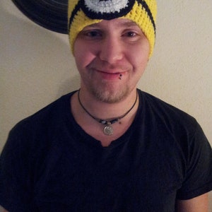 May include: A person wearing a yellow crocheted hat with a black and white eye design. The hat is pulled down over their forehead. The person is wearing a black t-shirt and a silver necklace.