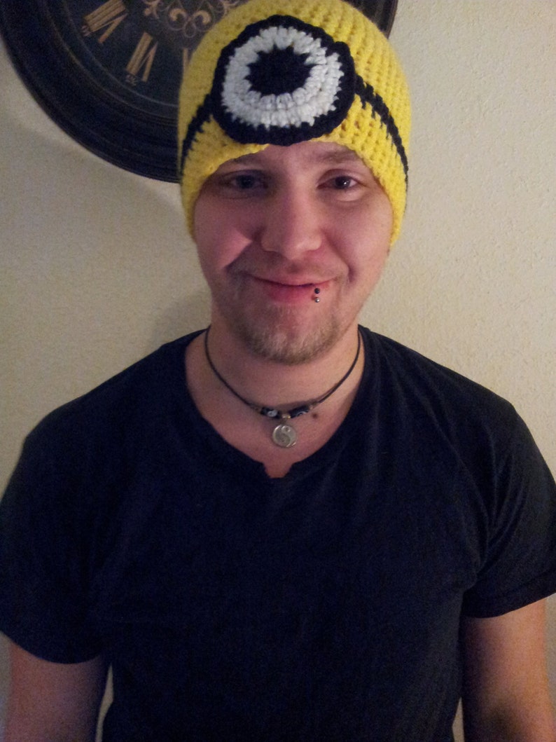May include: A person wearing a yellow crocheted hat with a black and white eye design. The hat is pulled down over their forehead. The person is wearing a black t-shirt and a silver necklace.