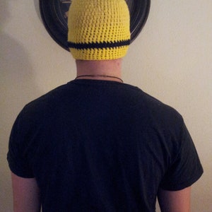 May include: A yellow crocheted beanie with a black stripe around the brim. The beanie is being worn by a person.