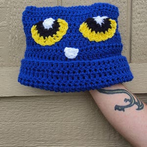 May include: A bright blue crochet beanie hat featuring a cat-like face design. The hat has two large yellow eyes with black pupils and white highlights, along with a small white nose detail centered below the eyes. The texture is a thick yarn knit.