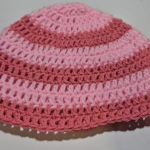 May include: A handmade crochet hat in shades of pink and rose. The hat features a striped pattern with alternating rows of light and darker pink yarn. The open weave design is visible throughout the hat.