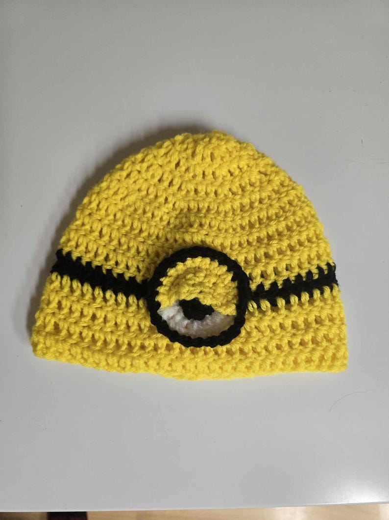 May include: A yellow crocheted hat with a black stripe and a single eye, resembling a Minion character.