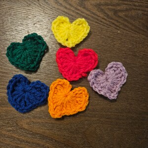 May include: Six crocheted hearts in different colors: yellow, green, red, blue, orange, and purple. The hearts are arranged in a semi-circle on a brown wooden surface.