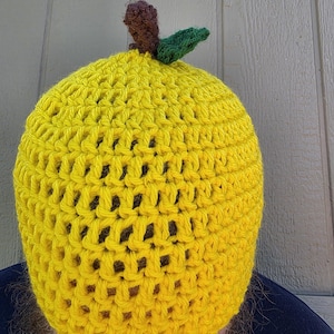 May include: A bright yellow crocheted beanie hat featuring a textured, open-stitch pattern. The top of the hat is adorned with a small, brown crocheted stem and a single, textured green leaf, mimicking the appearance of a piece of fruit.