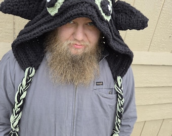 May include: A black crocheted hooded hat featuring two large, light green eyes with black pupils, pointed ears, and two long braided tassels made of black and light green yarn hanging down from the sides of the hood.