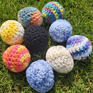 May include: A collection of nine colorful, crocheted balls of various sizes and hues, including yellow, blue, orange, and black. The balls are arranged on a bed of green grass, showcasing their handmade texture and vibrant colors.