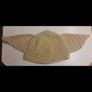 May include: A crocheted hat in a light tan color, featuring large, pointed ear-like extensions on either side. The hat is made with a visible stitch pattern, suggesting a handmade item. The design resembles a character from a popular science fiction franchise.