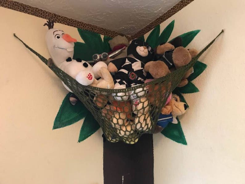 May include: A green net hammock with stuffed animals inside, including a snowman, a black and white striped animal, a brown animal, and a brown and white animal. The hammock is hanging from a wall painted to look like a tree with green leaves.